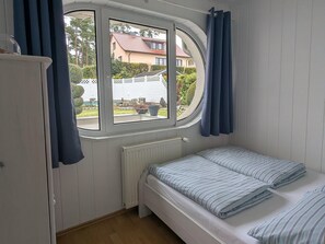A bedroom with a single bed, a window with curtains, a radiator, and a view of a house and garden. - Charmanter Bungalow in Neubukow Nahe der Ostsee (Neubukow)