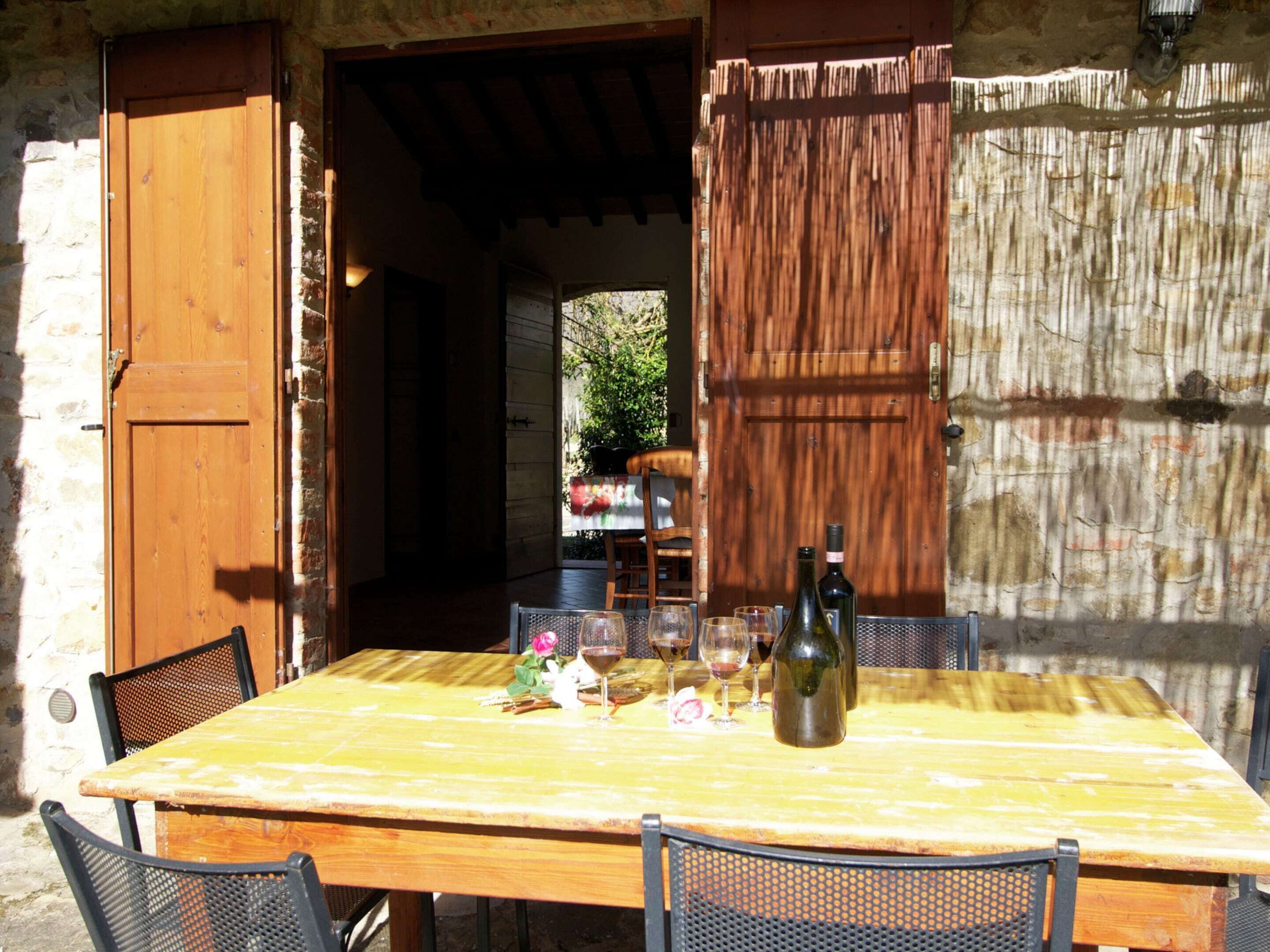 A rustic wooden table with a bottle and glasses, set against a stone wall with a wooden door and shutter.