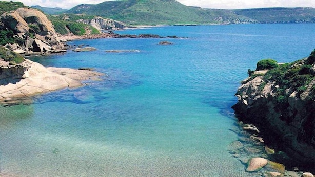 Paesaggio costiero con acque limpide, rocce a picco e colline verdi sullo sfondo.
