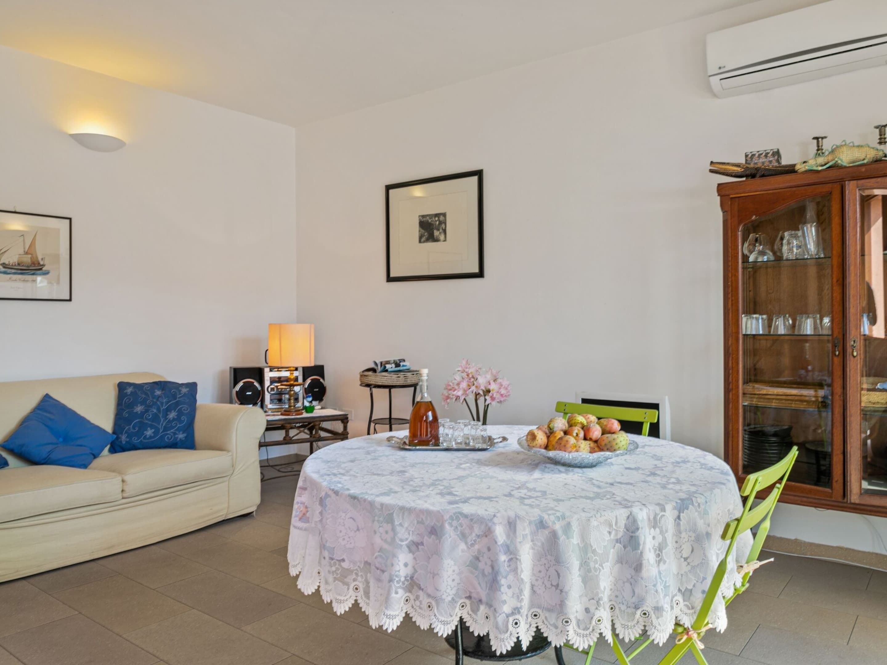 A living room with a round table covered in a lace tablecloth, a sofa with blue cushions, a small side table with a lamp, and a cabinet with glassware.