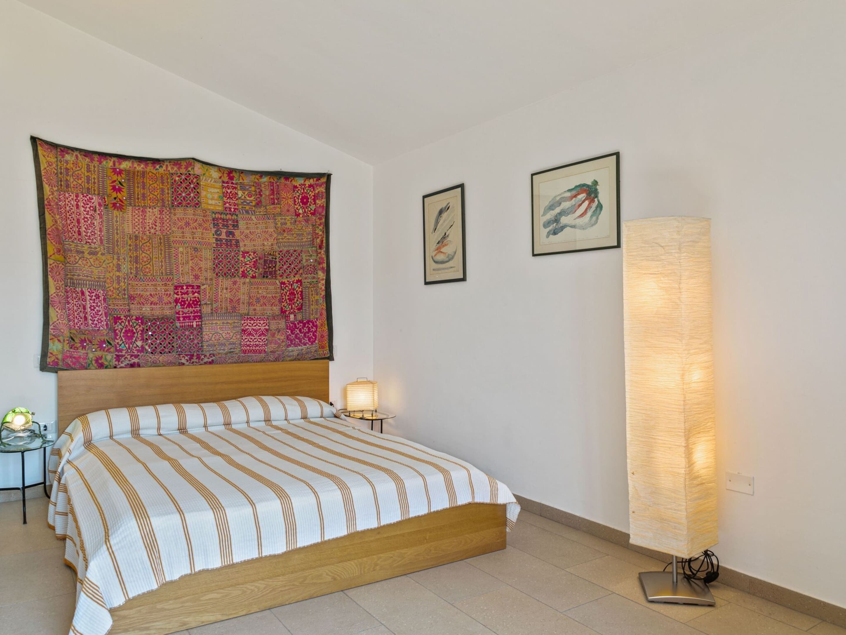 Une chambre à coucher avec un grand lit, une tête de lit, une tapisserie murale, deux tableaux encadrés, une lampe de chevet et une lampe sur pied.