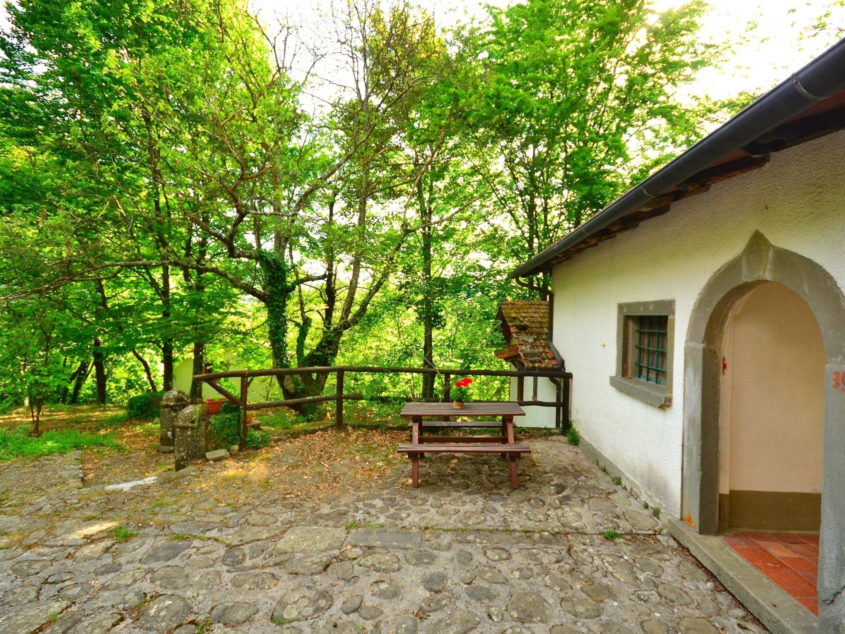 A rustic stone patio with a wooden bench and a table, surrounded by lush green trees. A white building with an arched entrance is visible.