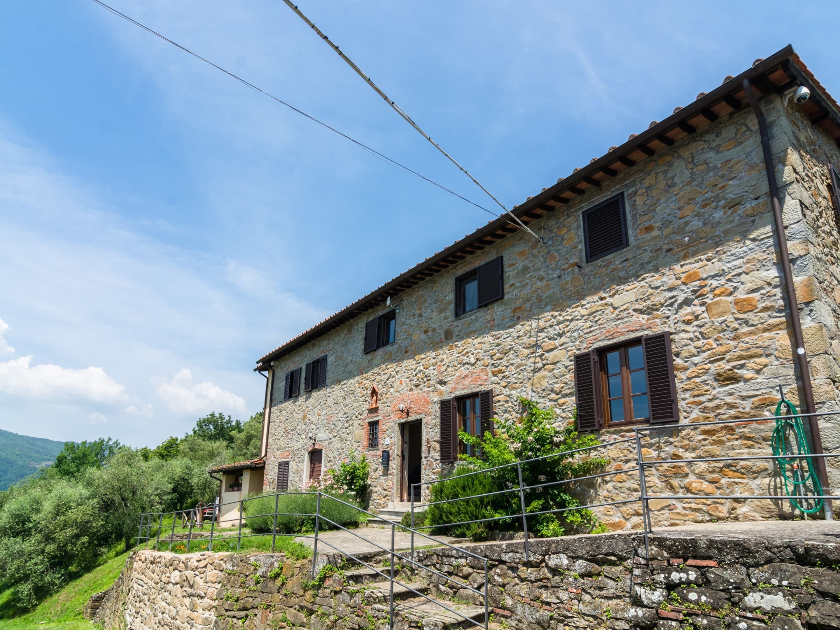 A stone building with multiple windows and a door, surrounded by greenery and hills.
