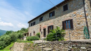 A stone building with multiple windows and a door, surrounded by greenery and hills.