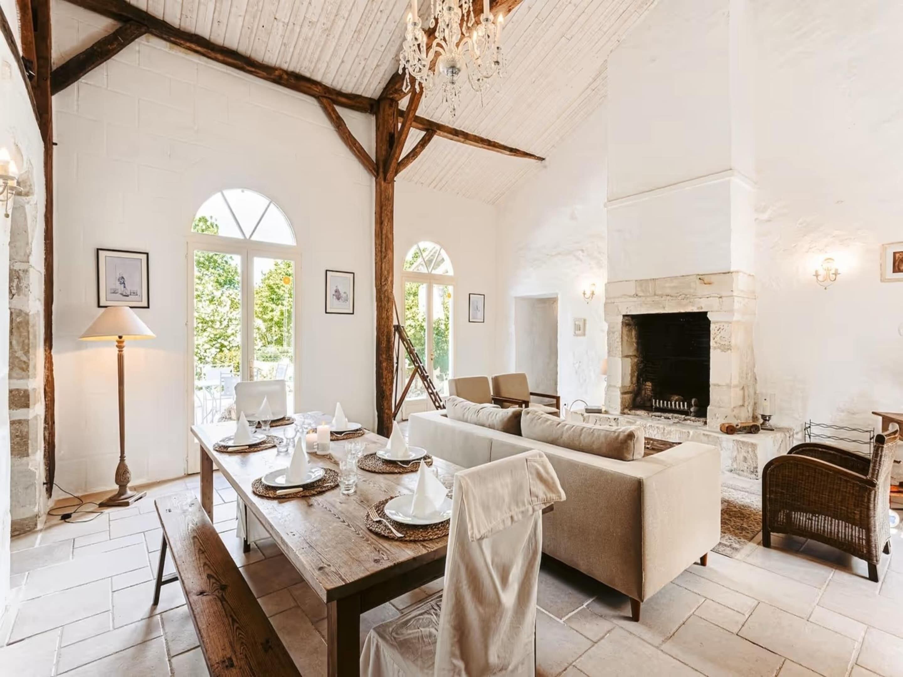 A spacious room with a wooden dining table set for four, a sofa, a fireplace, and a large window with a view of greenery.