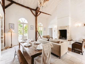 A spacious room with a wooden dining table set for four, a sofa, a fireplace, and a large window with a view of greenery. - Holiday Home Saint-laurent Pool & Garden (Saint-Laurent-de-la-Salle)