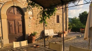 Zona exterior coberta amb taula i cadires, paret de pedra i plantes en testos.