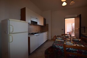 A compact kitchen with a refrigerator, stove, and cabinets. There is a dining table with chairs and a floral tablecloth. - Residence With Pool, Near the Beach and Coastal Town of La Ciacca (Valledoria)