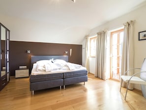 Quarto moderno com cama grande, piso de madeira, mesa de cabeceira, cadeira e janela com cortinas.