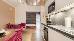 A compact kitchen with a microwave, coffee maker, and oven. There is a dining area with a wooden table and pink chairs. A bed is visible in the background. - Holiday Home in St. Georgen/salzburg (Bruck an der Grossglocknerstrasse)