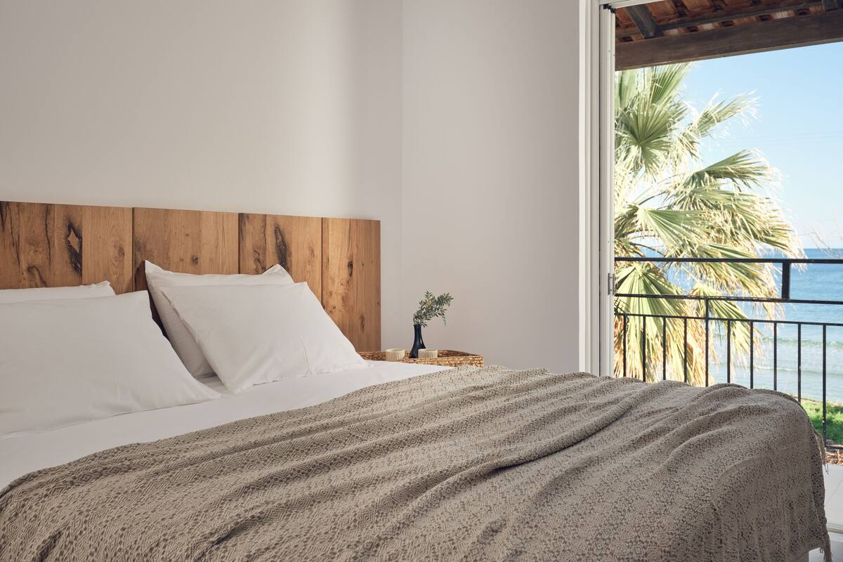 Une chambre à coucher avec une tête de lit en bois, du linge de lit blanc et une vue sur l’océan, visible à travers la rambarde du balcon.