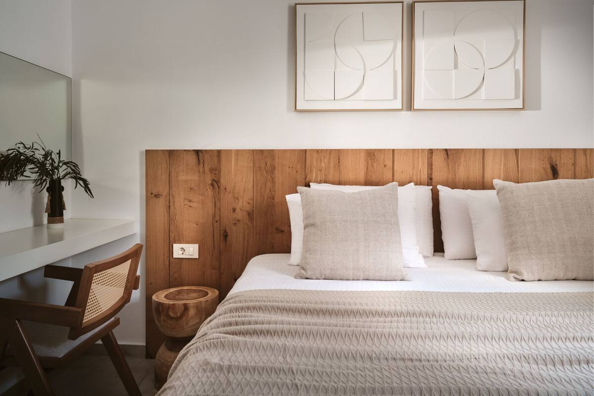Une chambre moderne avec une tête de lit en bois, un lit agrémenté de plusieurs coussins, une table de chevet, une chaise et deux tableaux abstraits accrochés au mur.
