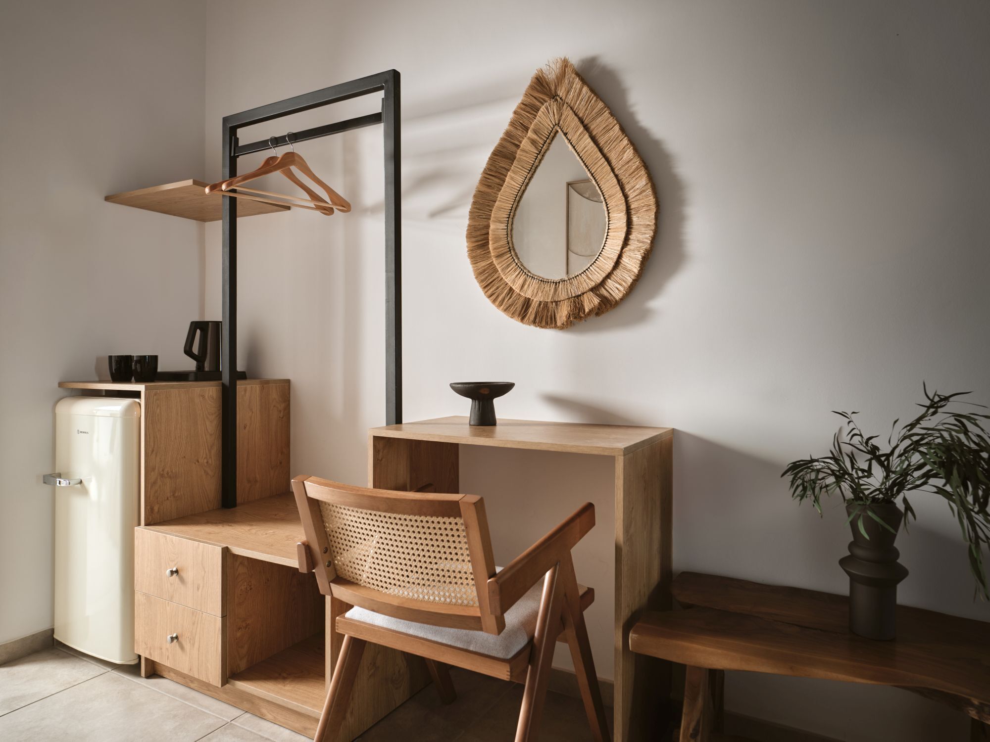 A minimalist room with a wooden desk, a chair, a hanging mirror, a small plant, and a coffee machine.