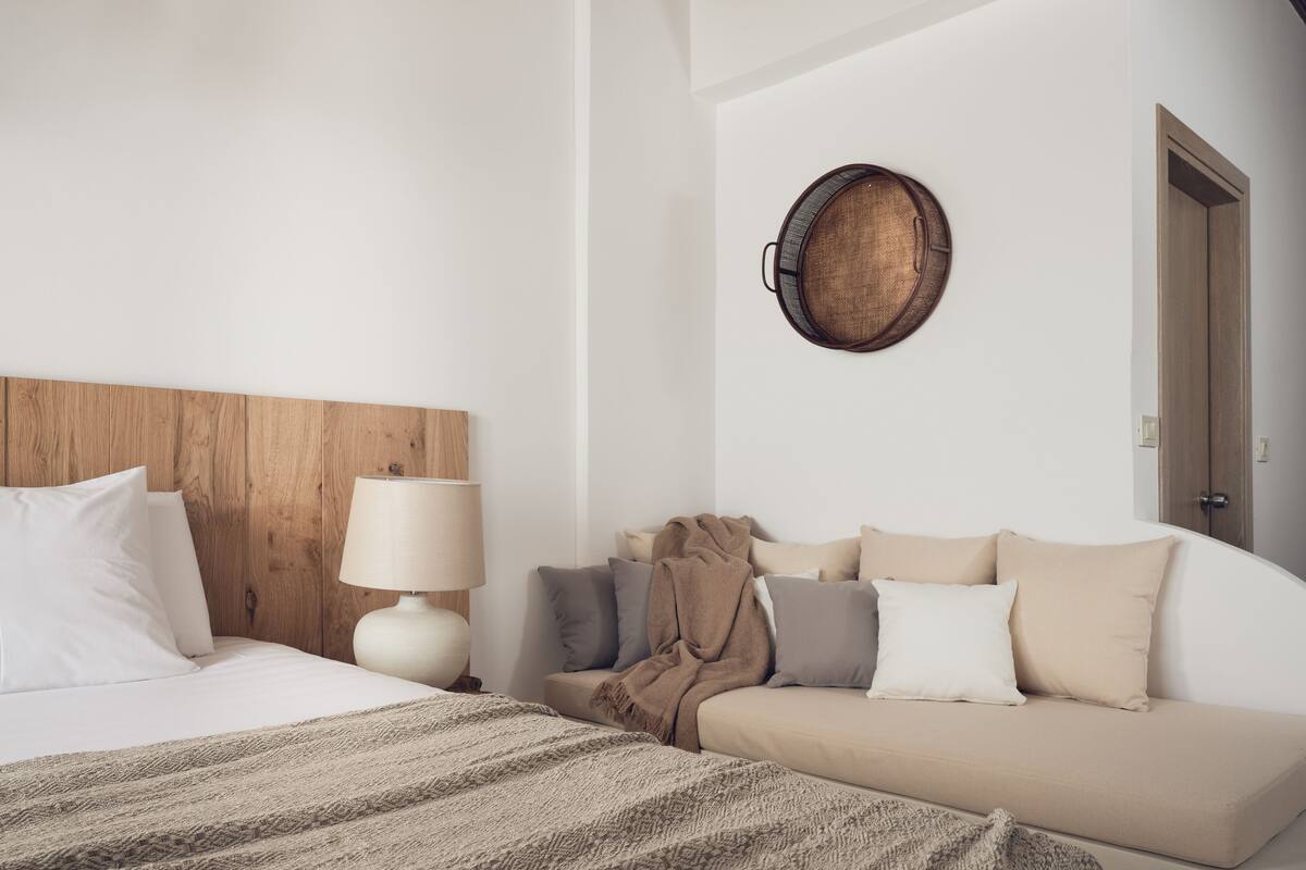 Une chambre d’hôtel moderne avec une tête de lit en bois, un panier rond en osier accroché au mur, un lit avec du linge de maison blanc, un canapé beige agrémenté de plusieurs coussins et une lampe de chevet.