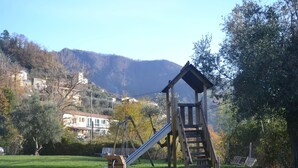 Un parque infantil con un tobogán y columpios, rodeado de un campo cubierto de césped y árboles, con casas y montañas al fondo.