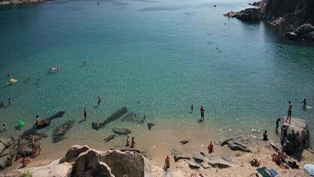 Une plage avec une eau claire, un rivage rocheux, et des gens qui se baignent et prennent des bains de soleil.
