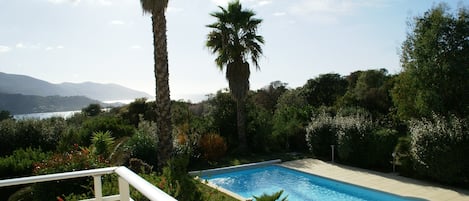 A swimming pool surrounded by lush greenery and palm trees, with a view of distant mountains and a body of water.