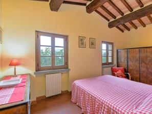 A bedroom with a checkered bedspread, a radiator, a window with a view, a wooden wardrobe, and a table with a lamp. - Scenic Stay in Restored Retreat (Monterchi)