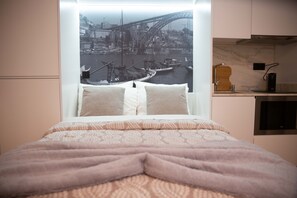 A bed with a view of a cityscape through a window, a bedside table with a lamp, and a small kitchen area with a microwave.
