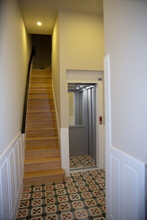 A narrow hallway with a wooden staircase leading to an elevator. The floor has patterned tiles.