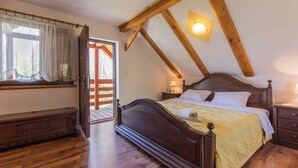 Un dormitori amb un llit de fusta, una finestra amb cortines, un paviment de fusta i un sostre inclinat amb bigues al descobert.