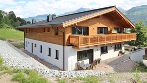 Une maison en bois à deux étages, dotée d’un balcon, qui se dresse sur un fond de paysage montagneux.
