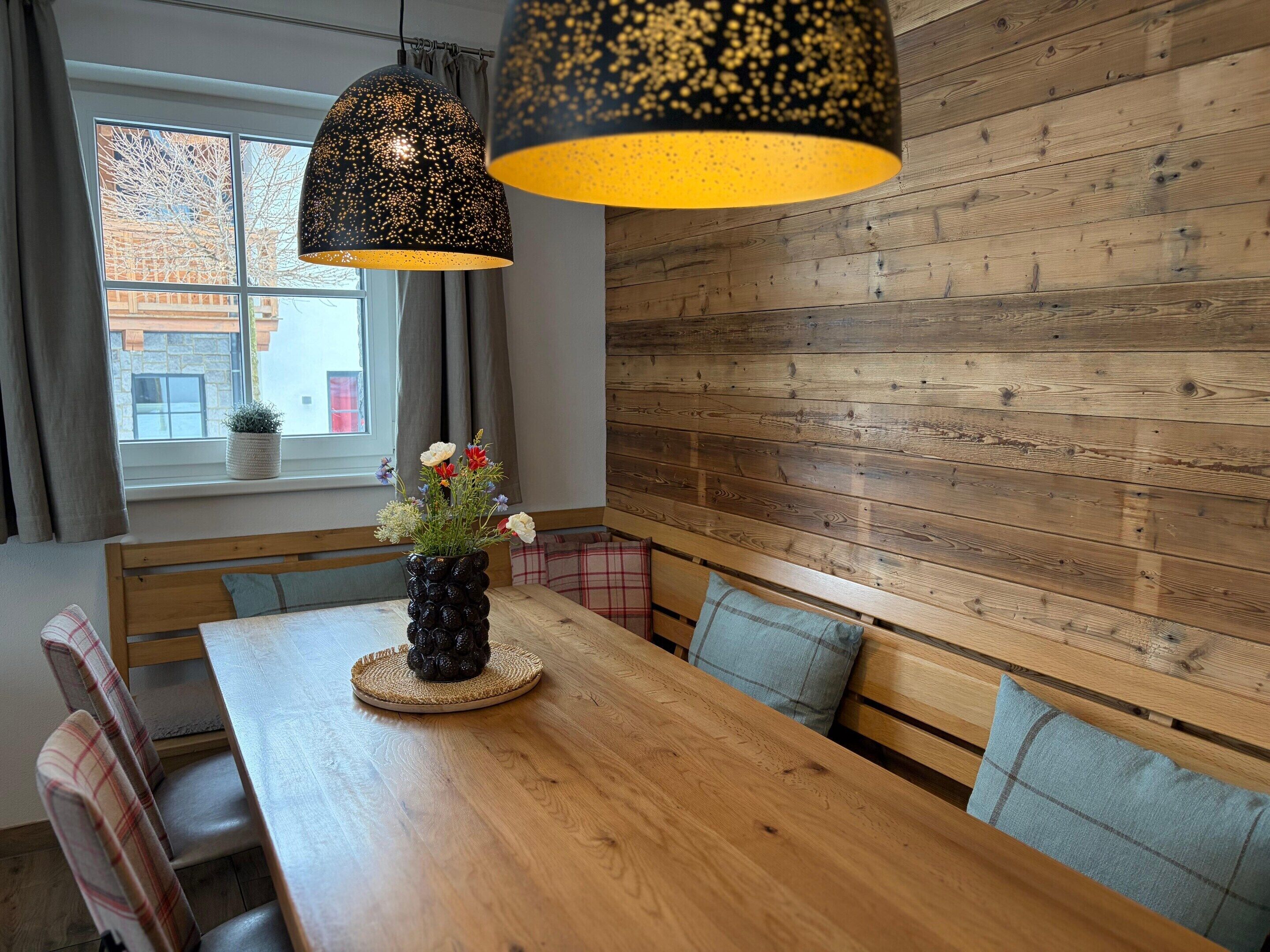 Une table à manger en bois, ornée d’un vase de fleurs, un banc avec des coussins et une fenêtre avec des rideaux.