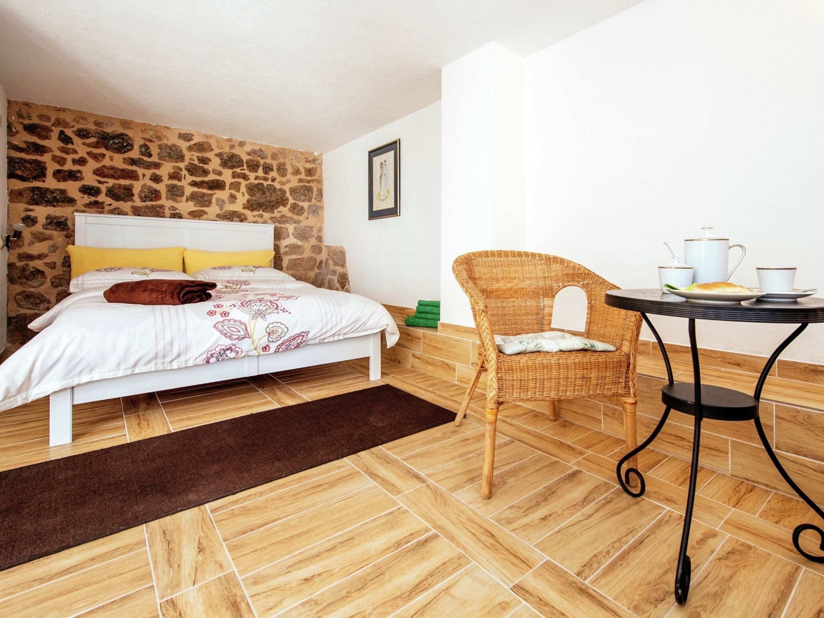 Een slaapkamer met een stenen muur, een bed met bloemenbeddelsels, een rieten stoel, een tafeltje met een theepot en kopjes, en een bruin vloerkleed.