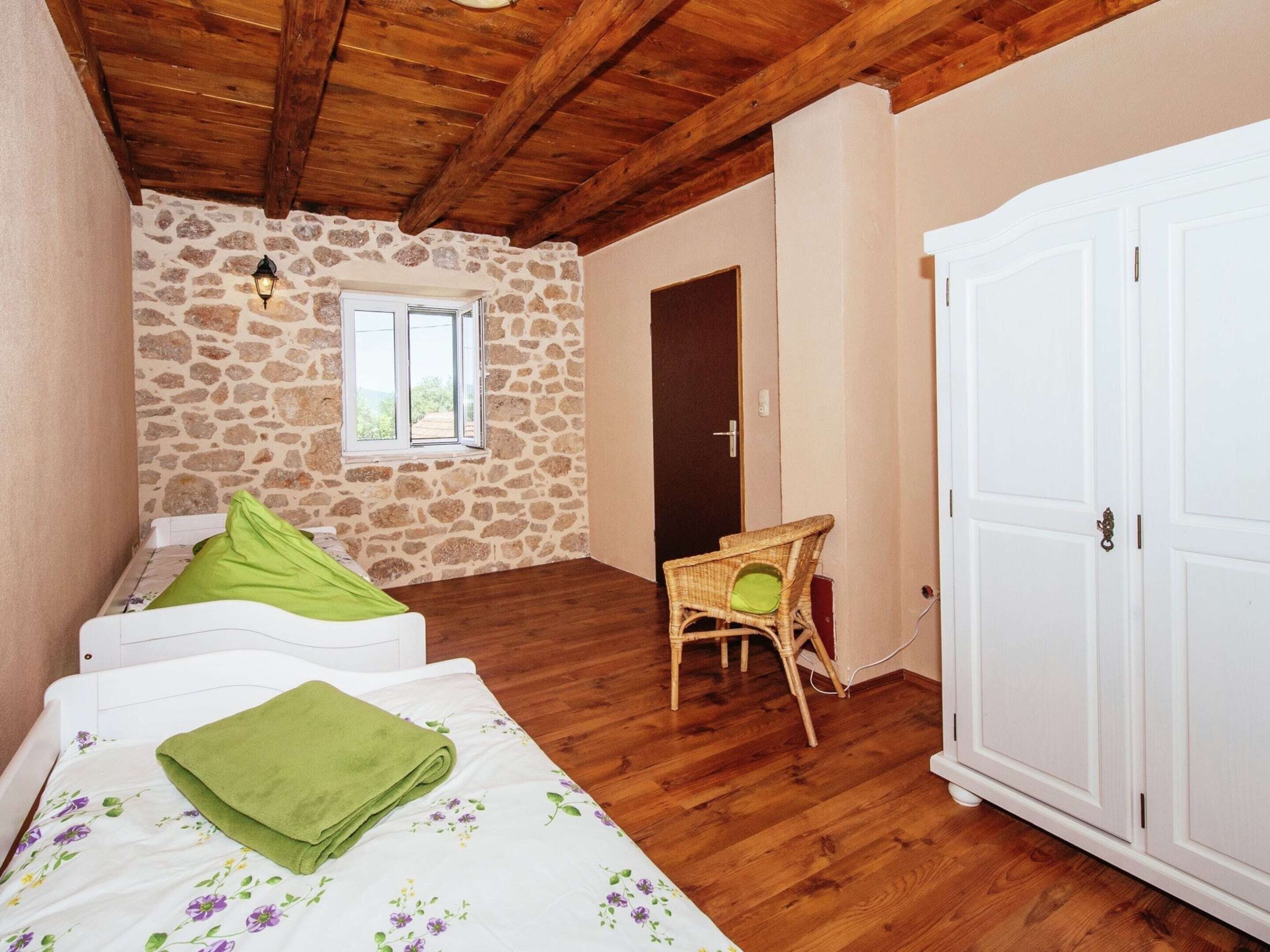 A bedroom with a stone wall, wooden ceiling, a bed with green bedding, a wicker chair, and a white wardrobe.