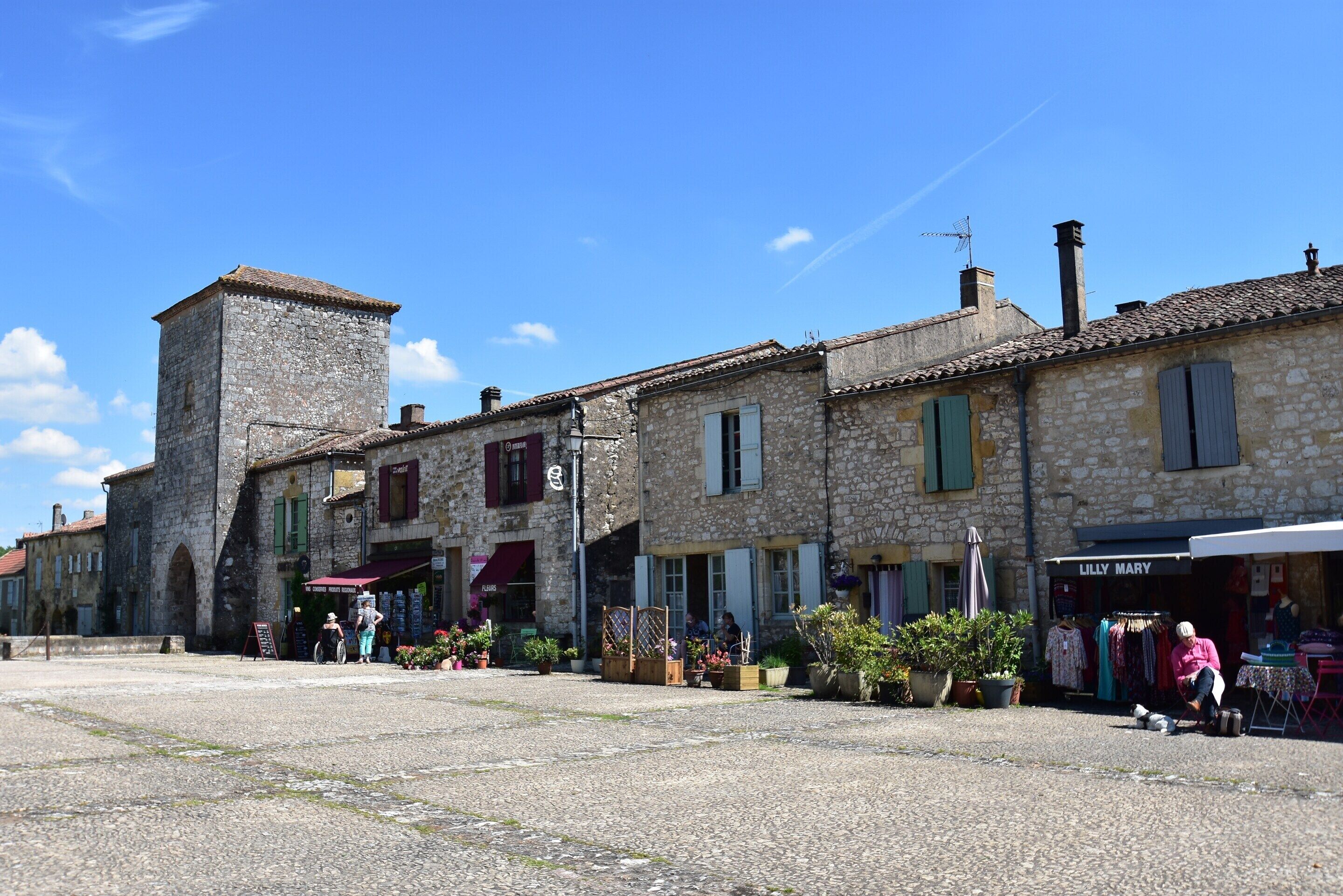 A stone building with a tower, a shop named 'Lilly Mary,' and outdoor seating with plants.