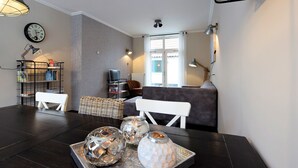 Ein modernes Wohnzimmer mit dunklem Holzboden, einem weißen Sofa, einem Esstisch mit dekorativem Glas und einem Bücherregal.