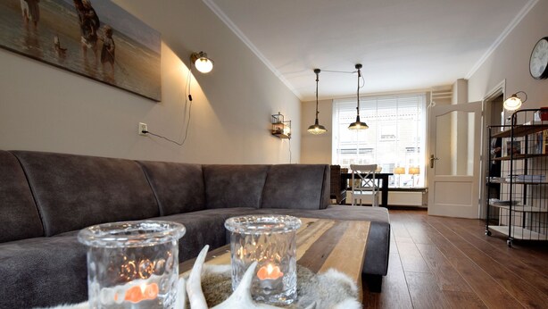 Ein modernes Wohnzimmer mit einem dunkelbraunen Sofa, einem Couchtisch aus Holz mit zwei Glaswindlichtern und einer weißen Felldecke. Über dem Sofa hängen Pendelleuchten, und rechts ist ein Bücherregal zu sehen.