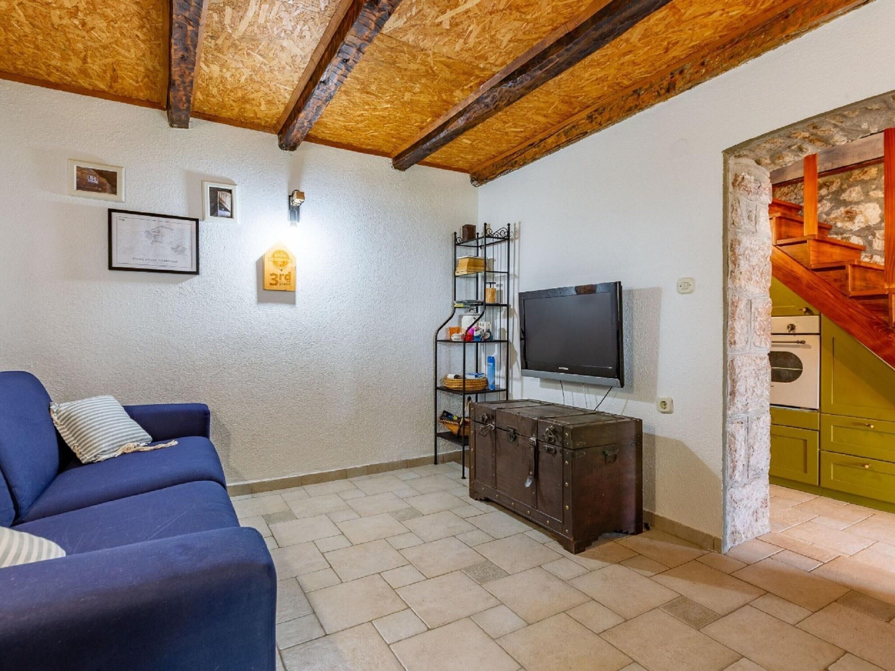A living room with a blue sofa, a wooden ceiling, a television, a suitcase, and a staircase leading to a kitchen.