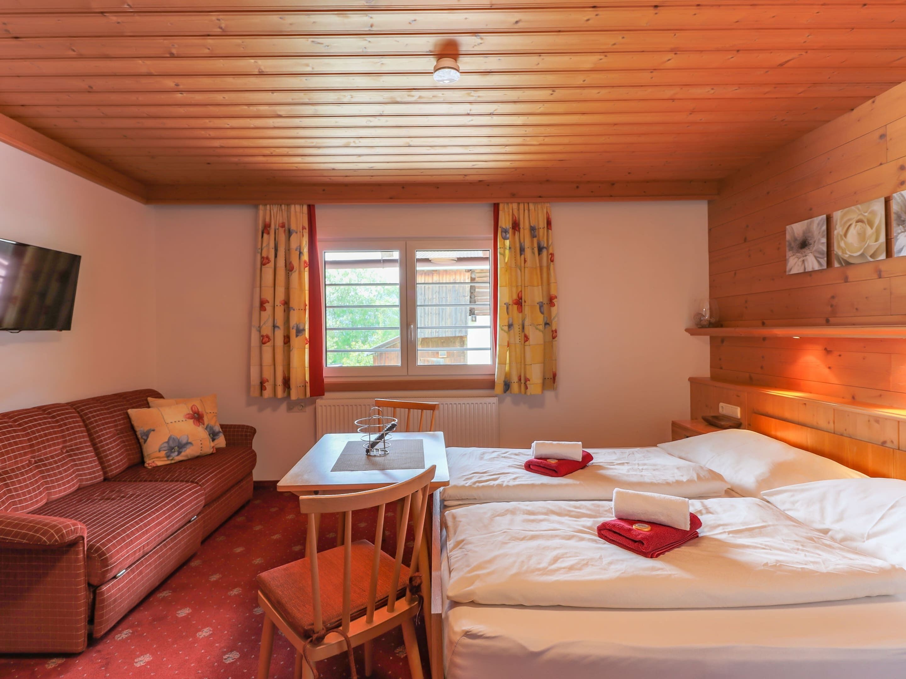 Ein gemütliches Zimmer mit Holzbalkendecke, einer Couch, einem kleinen Tisch mit Stuhl und zwei Betten mit Handtüchern.
