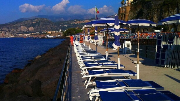 A seaside promenade with sun loungers and umbrellas, a rocky coastline, and buildings on a hillside. - Belvilla by OYO Casa Rosada (Pompei)