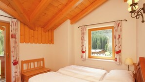Un dormitorio acogedor con techo de madera, un ventanal con cortinas, un banco de madera y una mesita de noche con lámpara.