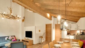 A modern living room with a wooden ceiling, a chandelier, a sofa, a TV, and a dining area set for six. - Luxurious Holiday Home With Sauna (Krimml)