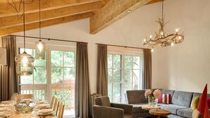 A spacious living room with a dining area, wooden beams on the ceiling, large windows, and a chandelier. - Luxurious Holiday Home With Sauna (Krimml)