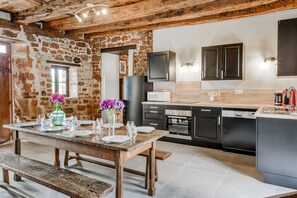 A rustic kitchen with stone walls, wooden beams, a wooden dining table set for four, and modern appliances. - Tranquil Holiday Home With Private Pool (Teillots)