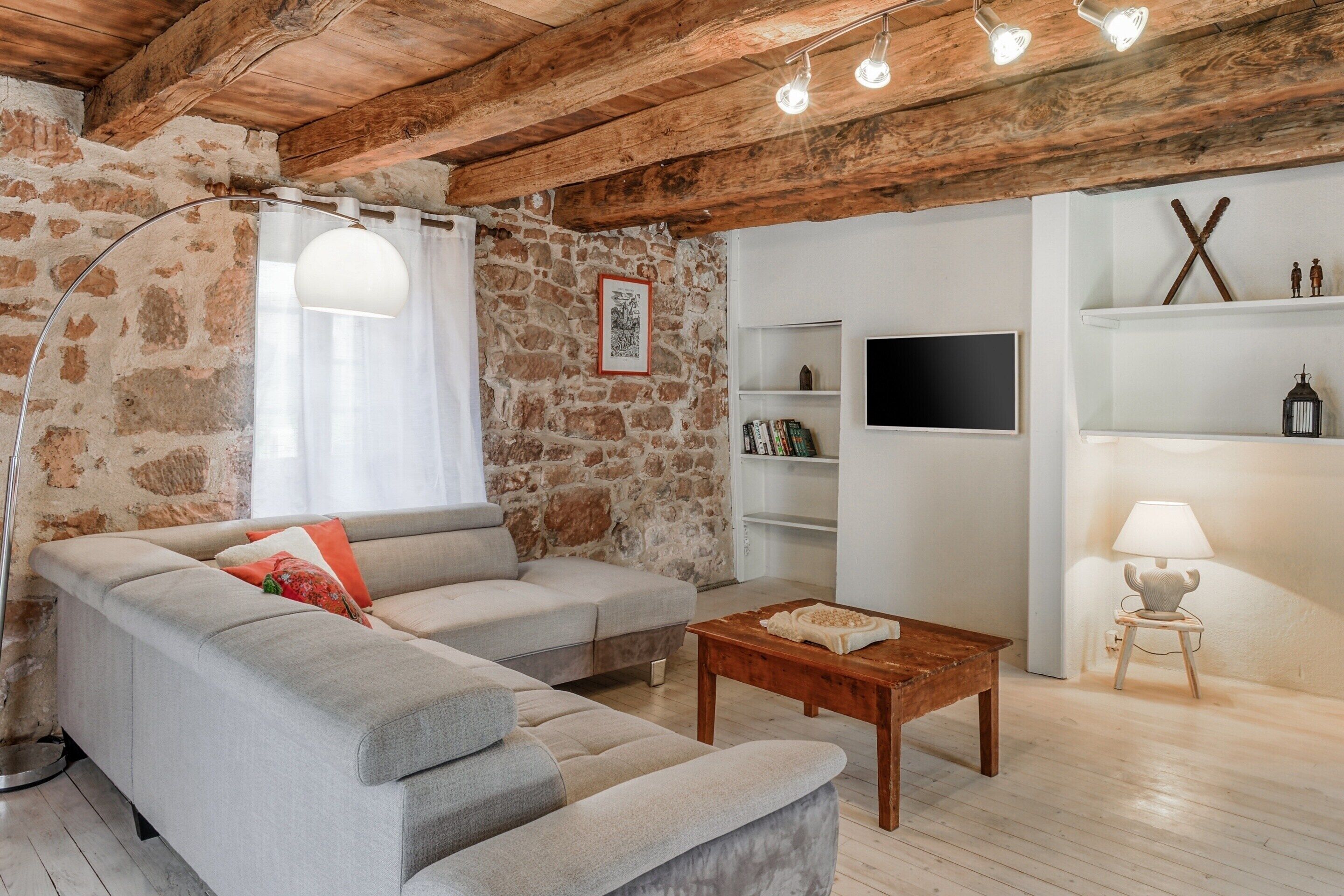 Un salon avec un mur en pierre, un canapé gris, une table basse en bois et un téléviseur posé sur une étagère blanche.