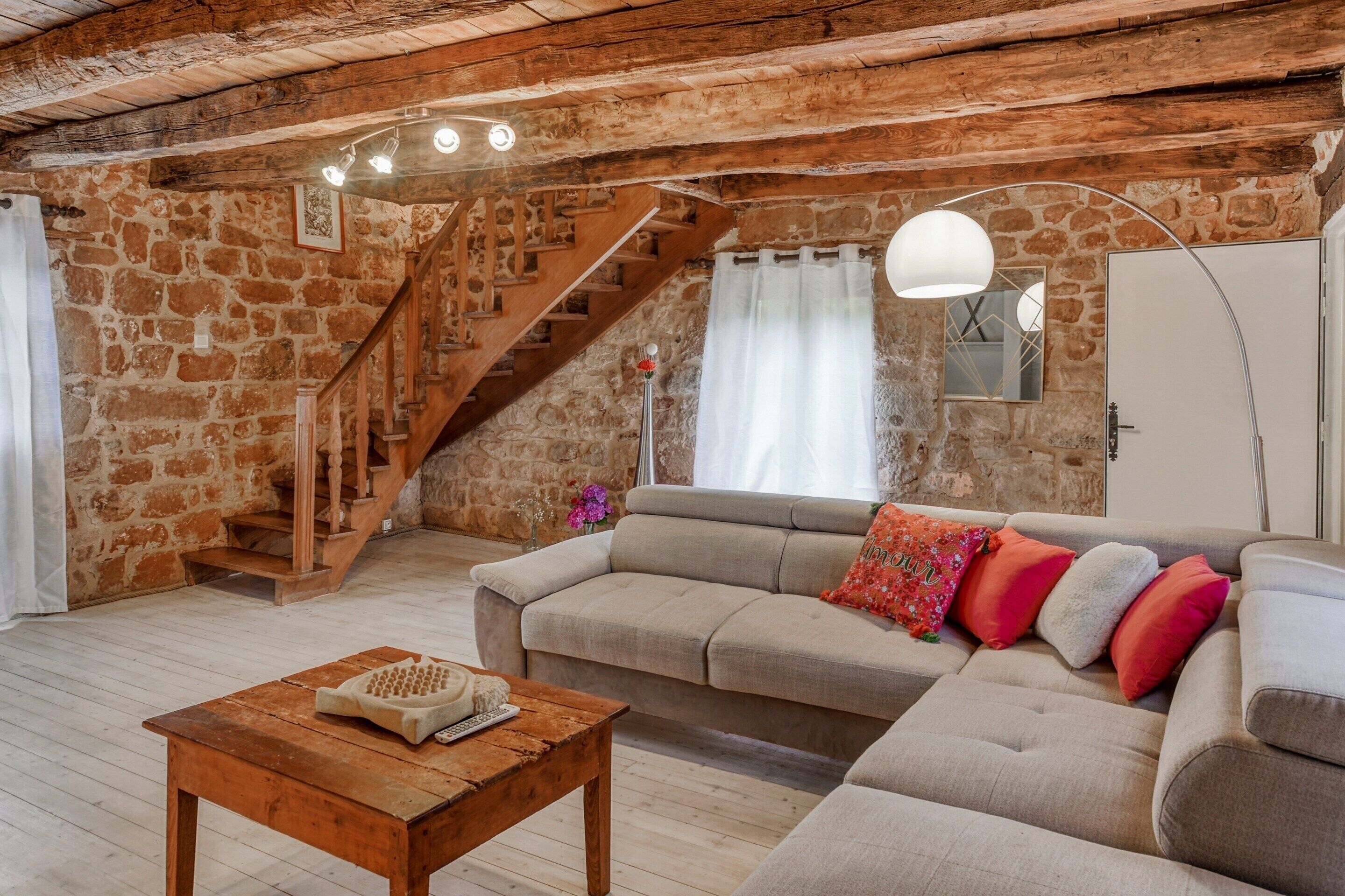 Una habitación rústica con paredes de piedra, una escalera de madera, un sofá color beige y una mesa de centro de madera.