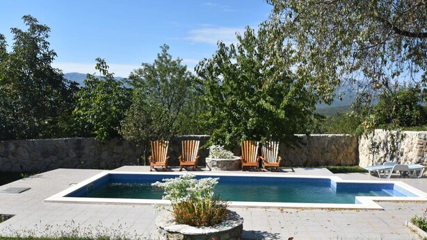 Une piscine rectangulaire bordée de pierres, entourée d’un muret en pierre. On y trouve des chaises en bois et une chaise longue disposées autour de la piscine.