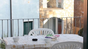 Varanda com mesa posta para dois, incluindo toalha de mesa com padrão náutico, duas cadeiras brancas e uma mesinha com uma tigela, um copo e uma xícara.
