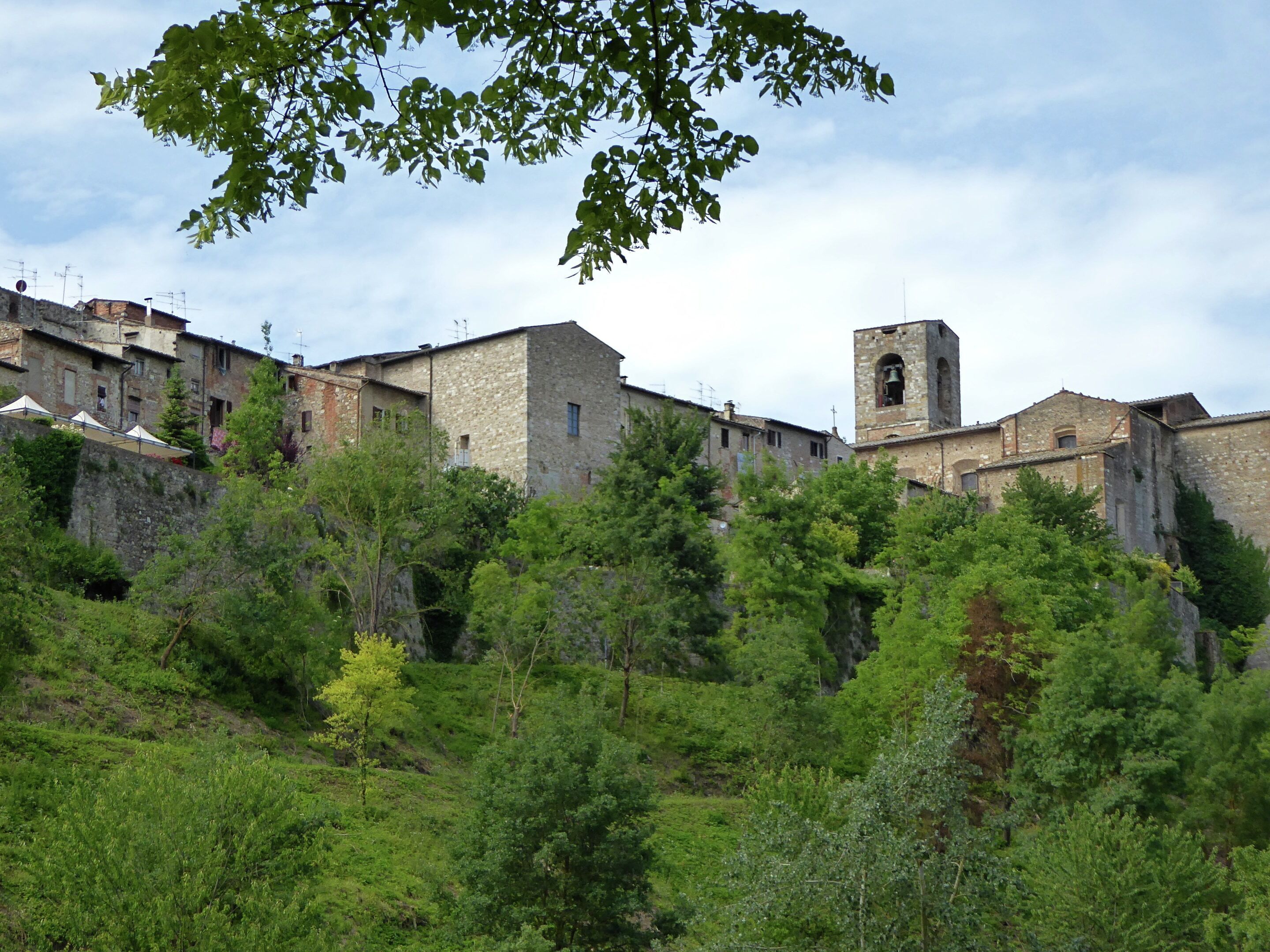 A historic stone building with a bell tower, surrounded by lush greenery and other stone structures.