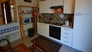 Una cucina compatta con mobili bianchi, microonde, piano cottura e lavastoviglie. Presenta una parete rivestita in piastrelle e un piccolo angolo ristoro con tavolo e sedie.