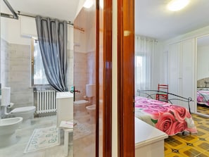 A bathroom with a shower, toilet, and sink. There is a bed with a floral bedspread and a chair. The room has white walls and a tiled floor. - Spacious Apartment near Saludecio (Saludecio)