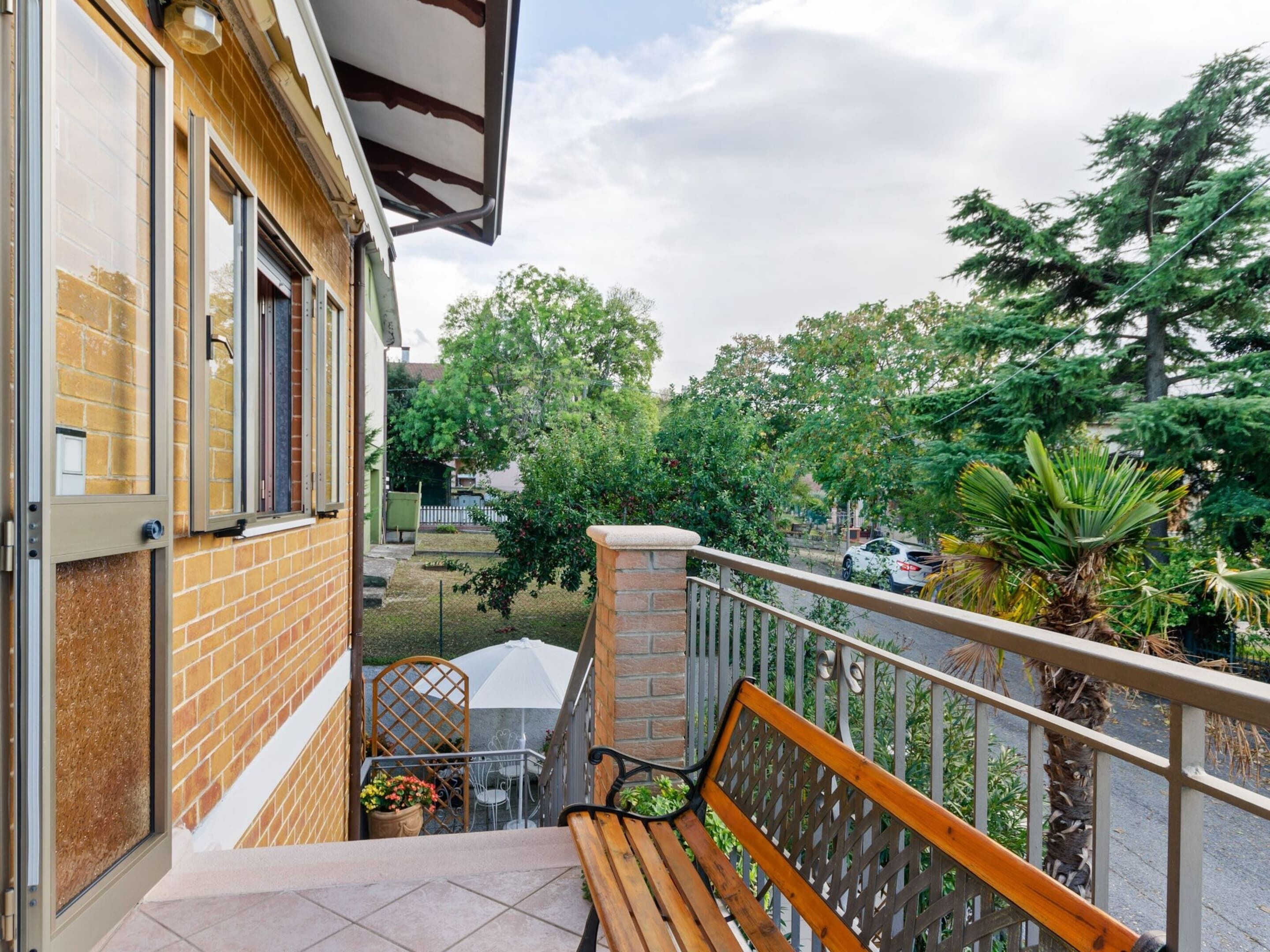 A balcony with a wooden bench, a potted plant, and a view of a street with cars and trees.