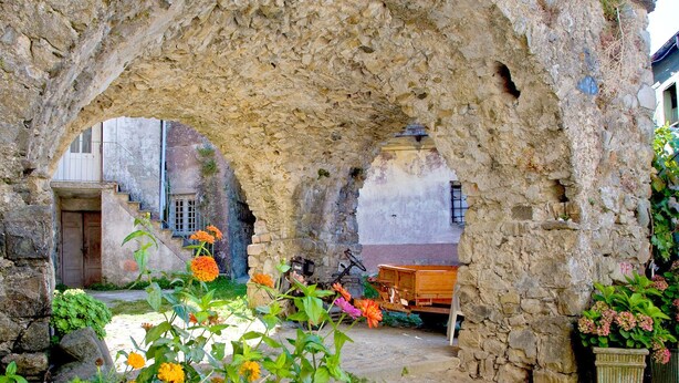 An ancient stone archway with a cobblestone courtyard, surrounded by flowering plants and a rustic wooden bench. - Apartment in Rio Near Roman Bridge (Sesta Godano)