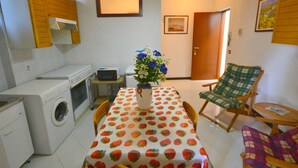A compact kitchen with a washing machine, a microwave, and a small table with a strawberry-patterned tablecloth. There is a chair with a plaid cushion and a small table with a single chair. - Apartment in Rio Near Roman Bridge (Sesta Godano)