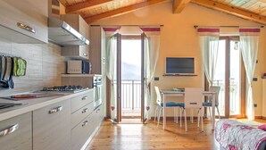 A modern kitchen with wooden cabinets, a gas stove, and a microwave. There is a dining area with a table and chairs, and a bed with a pink blanket. - Belvilla by OYO Romantica (Gravedona)